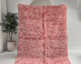 May include: A large, rectangular, blush-colored rug with a textured surface. The rug has a shaggy appearance and a fringe along the bottom edge. The rug is displayed in a bright, minimalist room setting.