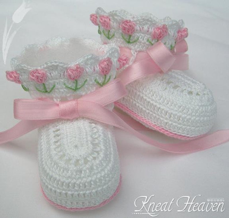 May include: A pair of white crocheted baby booties with pink satin ribbon ties and pink embroidered flowers.