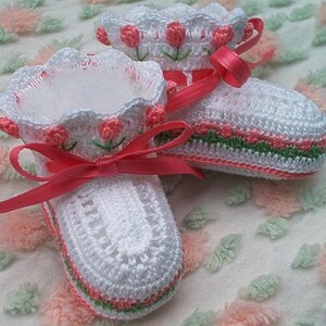 May include: A pair of white crocheted baby booties with pink and green trim. The booties have a pink satin ribbon tied around them.