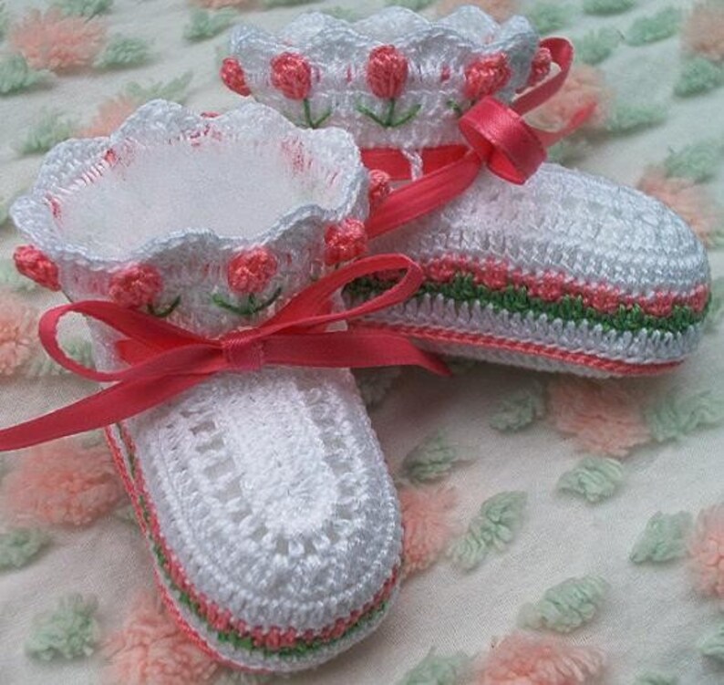 May include: A pair of white crocheted baby booties with pink and green trim. The booties have a pink satin ribbon tied around them.