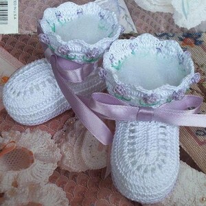 May include: A pair of white crocheted baby booties with a lavender ribbon tied around them. The booties have a delicate floral design and are lined with a soft white fabric.