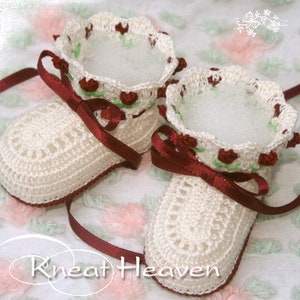 May include: A pair of white crocheted baby booties with burgundy satin ribbon ties and a floral design. The booties have a scalloped edge and are decorated with small red flowers and green leaves.