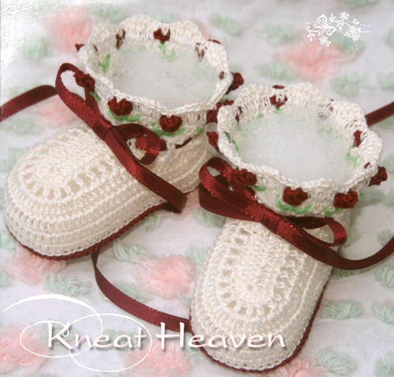 May include: A pair of white crocheted baby booties with burgundy satin ribbon ties and a floral design. The booties have a scalloped edge and are decorated with small red flowers and green leaves.
