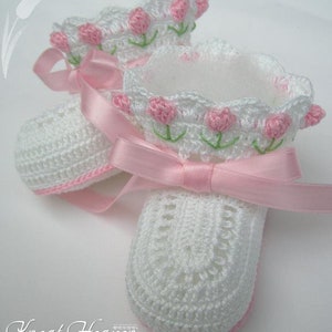 May include: White crocheted baby booties with pink ribbon ties and pink tulip accents. The booties have a scalloped edge and are lined with white fabric. The booties are on a white surface. The text 'Kneat Heaven' is visible in the bottom left corner of the image.