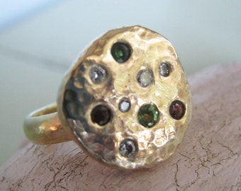 May include: A gold ring with a hammered round face set with multiple small gemstones in a variety of colors, including green, brown, and white.
