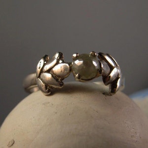 May include: A silver ring with a delicate leaf design and a single, round, grey diamond.