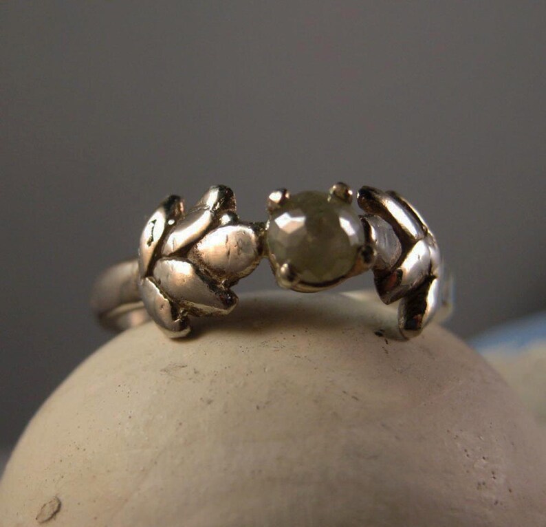 May include: A silver ring with a delicate leaf design and a single, round, grey diamond.