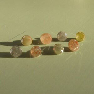 May include: A collection of eight small, round, faceted gemstones in various shades of brown, yellow, and gray. The gemstones are arranged in a semi-circle on a light green surface.
