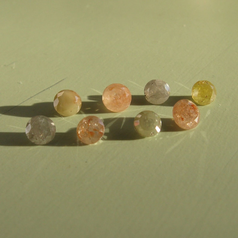 May include: A collection of eight small, round, faceted gemstones in various shades of brown, yellow, and gray. The gemstones are arranged in a semi-circle on a light green surface.