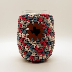 May include: A crocheted wine glass cozy with a brown leather patch featuring the outline of the state of Texas. The cozy is made with red, white, blue, and gray yarn.