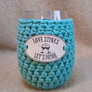 May include: A turquoise crocheted wine glass cozy with a white oval tag that reads "Love Stinks Let's Drink" with a graphic of two wine glasses clinking.