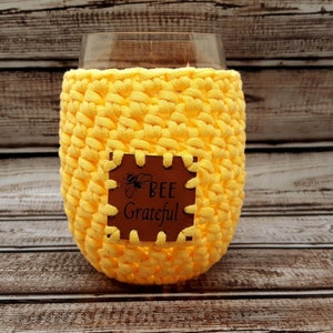 May include: Yellow crocheted wine glass cozy with a brown leather patch that says "Bee Grateful".