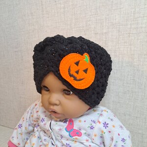 May include: A black crocheted baby hat with a glittery finish, adorned with an orange felt jack-o'-lantern applique. The baby is wearing a floral print onesie with a butterfly design.