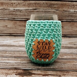 May include: A teal crocheted wine glass cozy with a cork patch that says "Stay Wild Ocean Child".