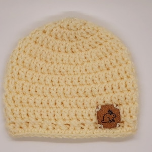May include: A crocheted beanie hat in a pale yellow color with a brown leather patch featuring a small rabbit design.