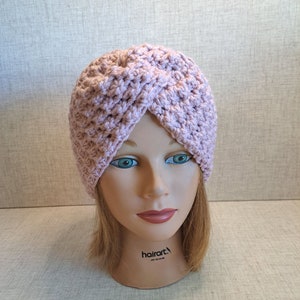 May include: A pink crocheted turban headband. The headband is on a mannequin head with brown hair.