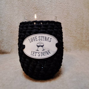 May include: Black crocheted wine glass cozy with a white oval tag that reads "Love Stinks Let's Drink" with a graphic of two wine glasses clinking.