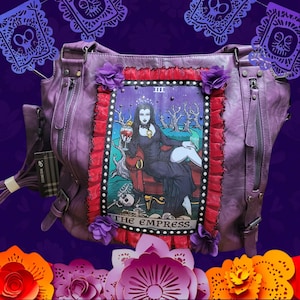 Puede incluir: Bolso de cuero morado con un diseño de carta de tarot que representa a la Emperatriz. La carta está rodeada de volantes rojos y blancos y flores moradas. El bolso tiene una cremallera y una correa.