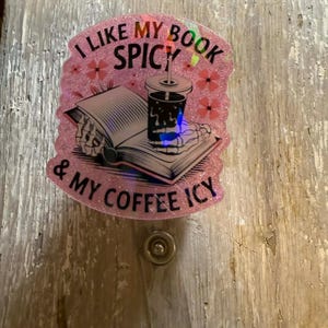 May include: A pink glittery badge reel with the text "I LIKE MY BOOK SPICY & MY COFFEE ICY." The design includes an open book, a coffee cup, and floral accents. The reel is attached to a silver button.