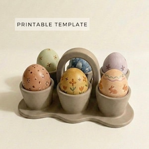 Op de afbeelding: Een grijze eierhouder voor Pasen met zes versierde eieren. De eieren zijn beschilderd met bloemen- en konijnenpatronen in pasteltinten. De houder heeft een handvat en de tekst "PRINTABLE TEMPLATE" is zichtbaar op de afbeelding.
