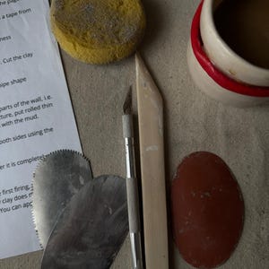 以下が含まれることがあります： 黄色いスポンジ、金属製の成形ツール、ナイフ、木製のツール、赤い縁取りのあるセラミックカップなど、陶器用のツールのコレクション。説明が書かれた紙も含まれています。ツールはニュートラルなトーンの表面に配置されています。