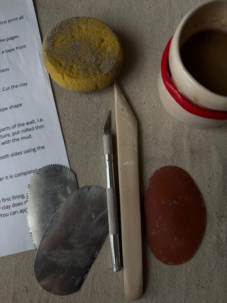 以下が含まれることがあります： 黄色いスポンジ、金属製の成形ツール、ナイフ、木製のツール、赤い縁取りのあるセラミックカップなど、陶器用のツールのコレクション。説明が書かれた紙も含まれています。ツールはニュートラルなトーンの表面に配置されています。