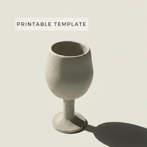 May include: A handmade, off-white ceramic wine glass with a textured surface. The glass has a rounded bowl, a slender stem, and a circular base. The words "PRINTABLE TEMPLATE" are printed in a white rectangle above the glass.