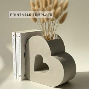 Op de afbeelding: Een hartvormige betonnen boekensteun met een kleinere hartvormige uitsparing. De boekensteun houdt drie boeken vast en is gevuld met gedroogd konijnenstaartgras. De tekst "PRINTABLE TEMPLATE" wordt weergegeven in een witte rechthoek.