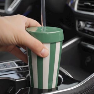 Puede incluir: Una taza de viaje a rayas verdes y blancas con una tapa a juego y una pajita transparente. La taza se sostiene en el portavasos de un coche. El dise&ntilde;o de la taza presenta rayas verticales verdes y blancas. La tapa es de color verde liso.