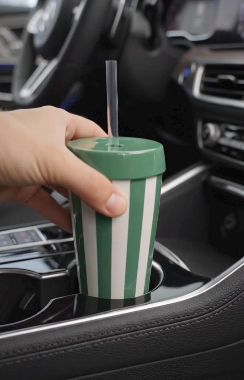 Puede incluir: Una taza de viaje a rayas verdes y blancas con una tapa a juego y una pajita transparente. La taza se sostiene en el portavasos de un coche. El dise&ntilde;o de la taza presenta rayas verticales verdes y blancas. La tapa es de color verde liso.