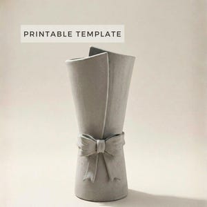 Op de afbeelding: Een grijze vaas met een strikdetail. De vaas heeft een unieke vorm, met twee naar buiten gebogen secties. De strik bevindt zich in de buurt van de basis van de vaas. De tekst "PRINTABLE TEMPLATE" staat bovenaan.