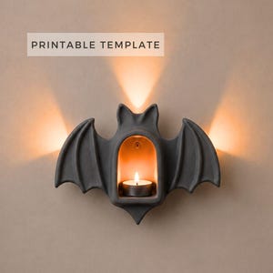 May include: A dark gray bat-shaped wall sconce featuring a central arched alcove that holds a small lit tea light candle. The design includes the text PRINTABLE TEMPLATE at the top. The sconce emits a warm glow from behind its wings and center.