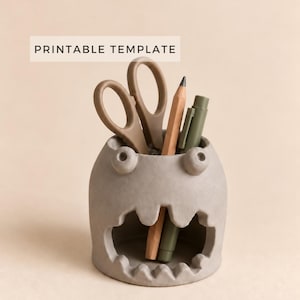 Puede incluir: Un portalápices gris con forma de monstruo, con una boca ancha y dentada y dos ojos pequeños en la parte superior. El soporte contiene unas tijeras marrones, un lápiz de madera y un rotulador verde. El texto PRINTABLE TEMPLATE aparece arriba.