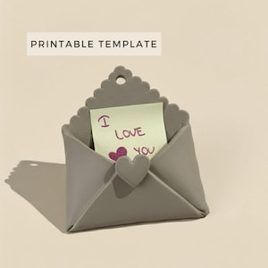 以下が含まれることがあります： 波状のトップとハート型の留め金が付いた、灰色の封筒型のホルダー。内側には、ハートのシンボルが付いた「I Love You」と書かれた黄色の付箋があります。「PRINTABLE TEMPLATE」というテキストが上部にあります。