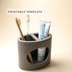 May include: A gray concrete toothbrush holder with a rounded design, holding a bamboo toothbrush, a clear toothbrush with rainbow bristles, and a tube of toothpaste. The text "PRINTABLE TEMPLATE" is displayed at the top.