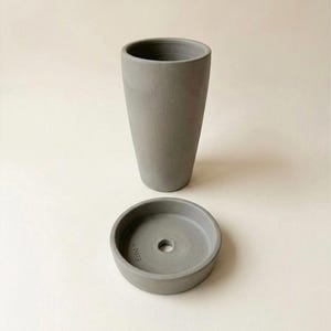 Puede incluir: Un juego de maceta y platillo de hormig&oacute;n gris. La maceta es alta y cil&iacute;ndrica, mientras que el platillo es redondo con un orificio de drenaje central. El juego est&aacute; dise&ntilde;ado para uso en interiores o exteriores.