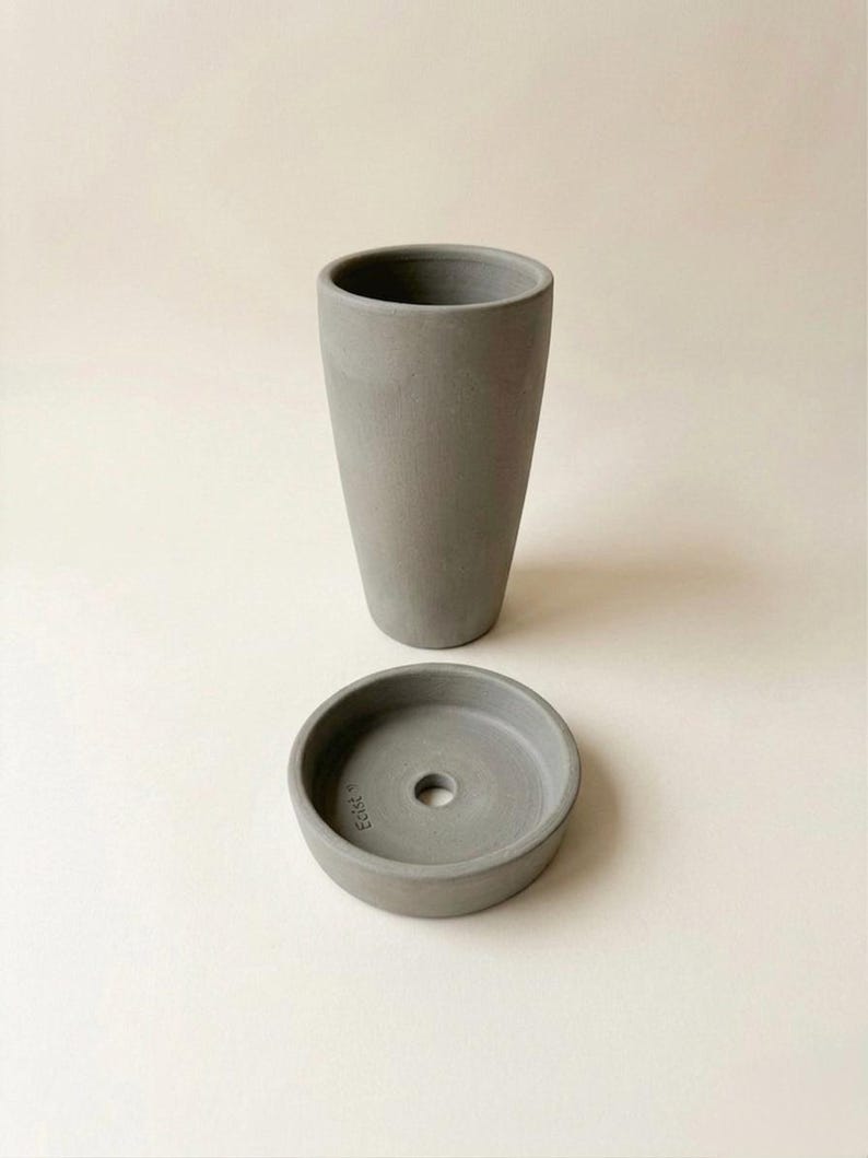 Puede incluir: Un juego de maceta y platillo de hormig&oacute;n gris. La maceta es alta y cil&iacute;ndrica, mientras que el platillo es redondo con un orificio de drenaje central. El juego est&aacute; dise&ntilde;ado para uso en interiores o exteriores.