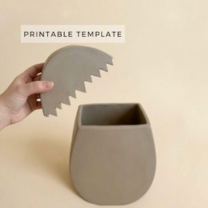 Puede incluir: Un jarrón gris moderno con una parte superior cuadrada y una base redondeada, y una tapa a juego con un borde dentado. El texto "PRINTABLE TEMPLATE" se muestra encima del jarrón.