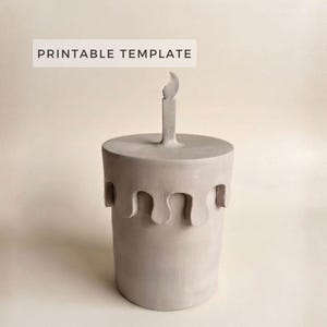Puede incluir: Un recipiente cilíndrico de cerámica gris claro con una tapa que presenta una vela y una llama esculpidas en la parte superior. La tapa tiene un borde festoneado decorativo que cuelga por los lados. El texto PRINTABLE TEMPLATE es visible arriba a la izquierda.
