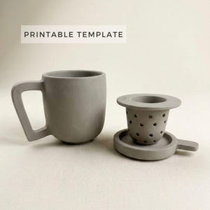 Può includere: Una tazza in ceramica grigia con un design unico del manico e un set di infusore per tè abbinato. L'infusore include un cestello forato e un piccolo vassoio. Il testo "PRINTABLE TEMPLATE" è visualizzato sopra la tazza.