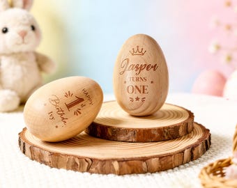May include: Two smooth, light-coloured wooden eggs engraved with text. One egg stands upright and reads "Jasper TURNS ONE" with a crown icon. The other egg lies on its side and reads "HAPPY 1st Birthday IVY" with star and sunburst accents.