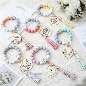 Puede incluir: Una colección de llaveros de cuentas de silicona coloridas con detalles de madera y borlas. Cada llavero presenta un dije de madera redondo con texto o diseños personalizados, incluidos nombres y la frase "best teacher ever."