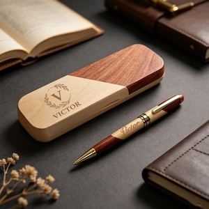 Può includere: Un set penna e astuccio in legno. La penna è in legno marrone e chiaro con dettagli dorati, e l'astuccio è bicolore con una base in legno chiaro e un coperchio in legno più scuro. Il nome "Victor" è inciso.