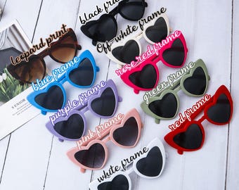 May include: A collection of heart-shaped sunglasses featuring various frame colors including black, off-white, rose red, blue, purple, green, red, pink, and white, along with a leopard print pair, all displayed with their respective color labels.