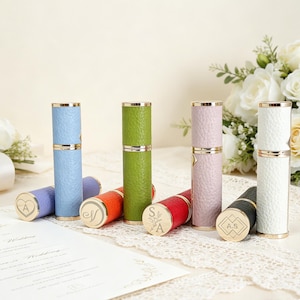 May include: A collection of cylindrical perfume atomizers with textured faux leather covers in light blue, green, lavender, white, red, orange, and black. Each atomizer features gold-tone metal accents and a flat top engraved with initials or symbols.