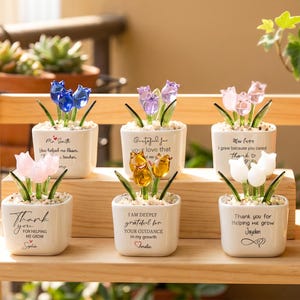 Peut inclure: Six petits pots carrés en céramique blanche contenant chacun trois fleurs de tulipes en verre bleu, violet, rose, ambre ou blanc. Chaque pot présente un texte imprimé en noir exprimant de la gratitude, comme Thank you for helping me grow.
