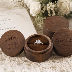 May include: Three round dark brown wooden ring boxes with lids. One box is open, revealing a silver ring with a clear stone set in brown velvet. The lids are engraved with mountain graphics and names including Emily & Daniel and Ryan & Kelly.