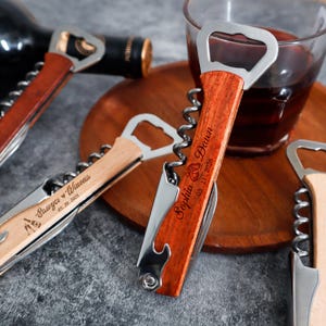 May include: A set of stainless steel wine openers with wooden handles. Each opener features a metal corkscrew, a bottle opener, and a small blade. The handles are engraved with names and dates, including 'Sawyer & Winona 05.20.2025' and 'Sophia & Dawn 03.11.2026'.