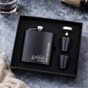 May include: A matte black stainless steel flask set presented in a black gift box. The flask features the engraved text Delilah, 10.24.26, and BRIDESMAIDS. The set includes two matching black shot cups and a small silver metal funnel for filling.
