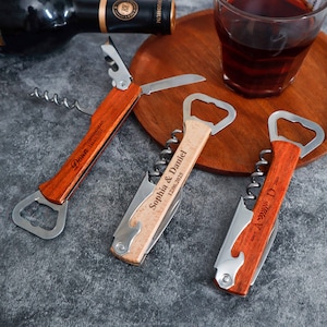 May include: Three multi-functional wine openers with wooden handles and stainless steel components. Each tool features a corkscrew, a bottle opener, and a small blade. The handles are engraved with names and dates, including Dawn, Sophia & Daniel, and A & D.
